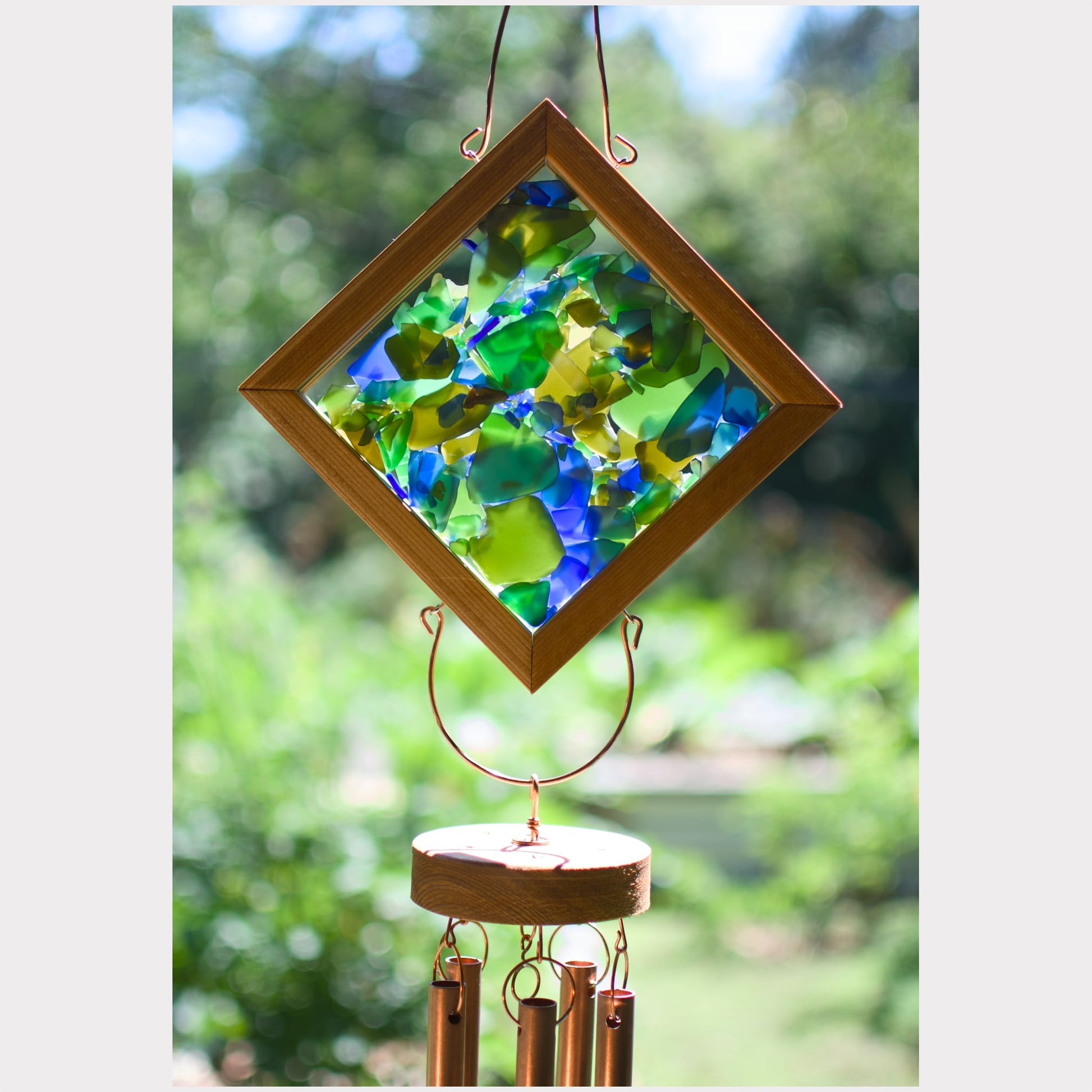 Diamond-shaped suncatcher with colorful stained glass panels in green, blue, and yellow, framed in wood and hanging from a wind chime outdoors.