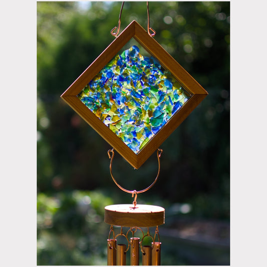 A diamond-shaped wind chime with a wooden frame and colorful broken glass insert, hanging outdoors against a blurred green garden background.