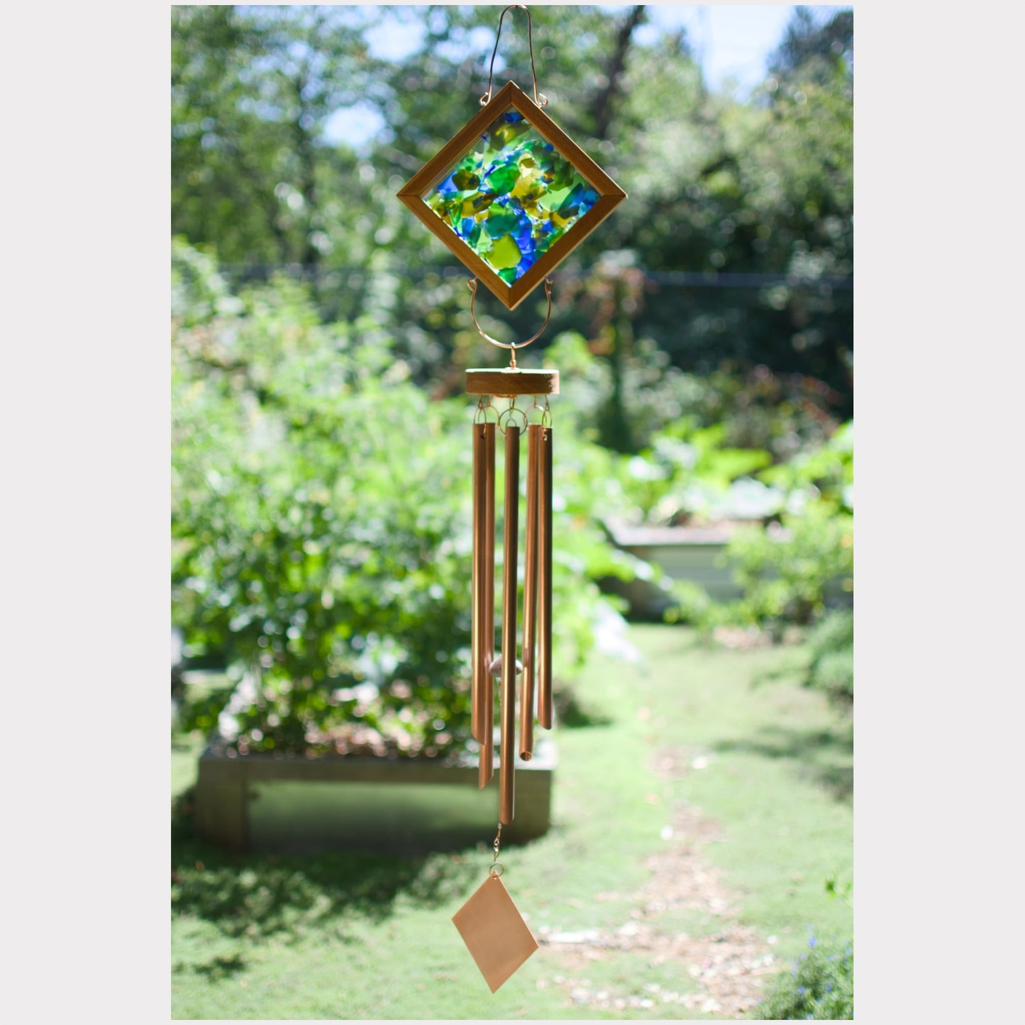 A wind chime hanging outdoors in a garden, featuring a diamond-shaped stained glass top with green, blue, and yellow leaves, copper-colored metal tubes, and a small hanging tag at the bottom.