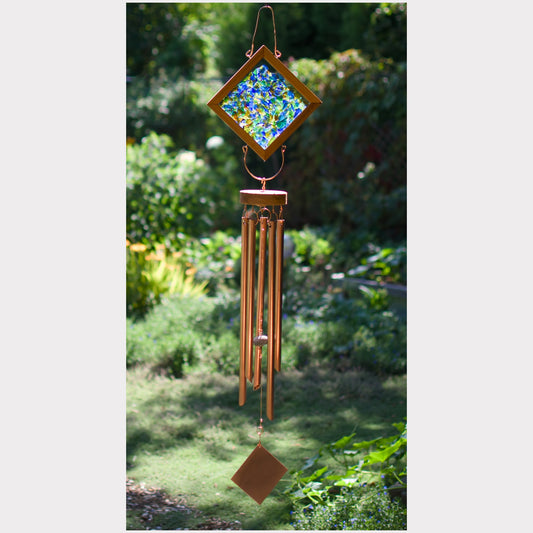 A wind chime hanging outdoors in a garden, featuring a diamond-shaped stained-glass top with blue, green, and yellow tones, copper-toned metal tubes, and a small hanging ornament at the bottom.