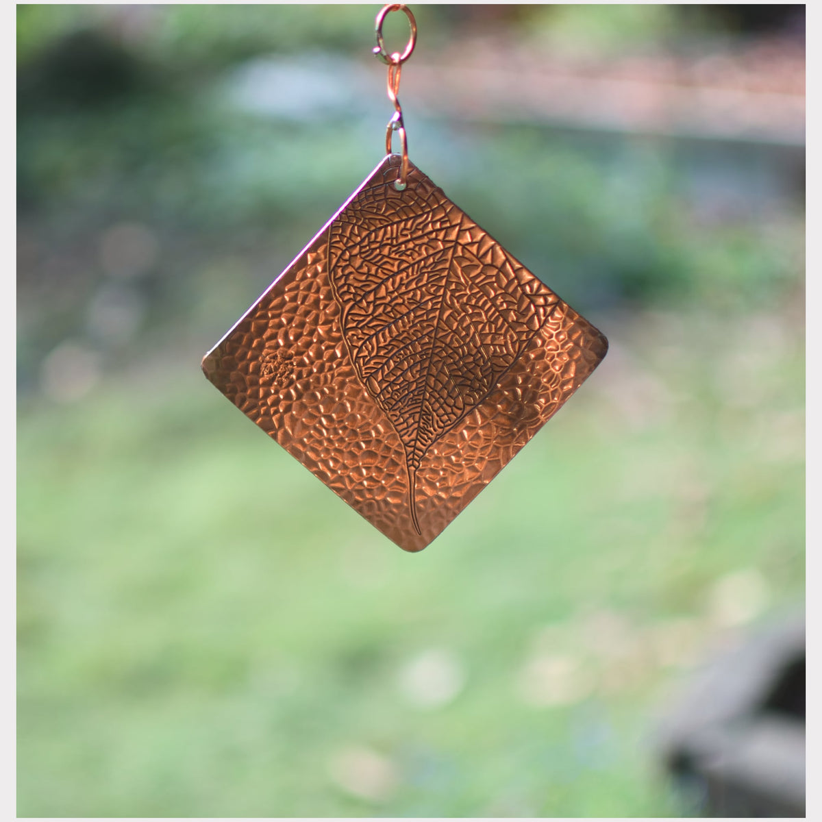 A hammered copper square pendant with a leaf design, hanging from a small copper loop against a blurred green outdoor background.